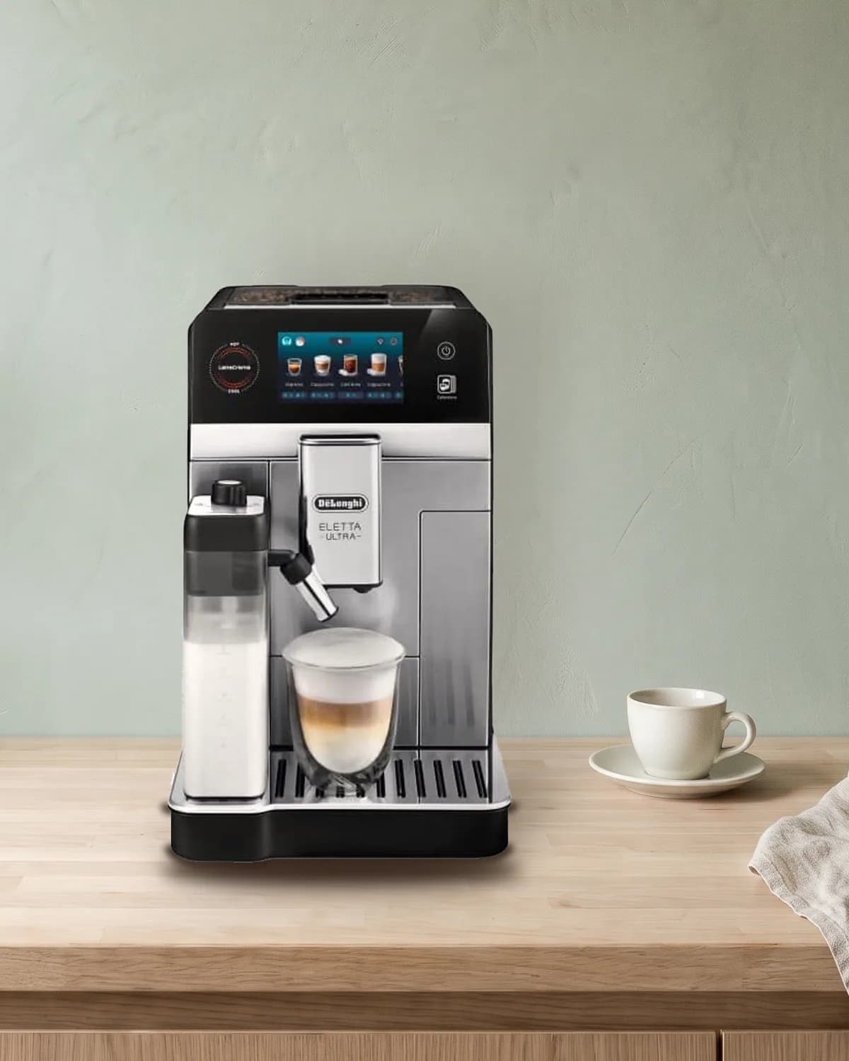 De'Longhi Eletta Ultra volautomaat in zilver en zwart met melkkan en latte macchiato op een licht eiken aanrecht tegen een saliegroene muur